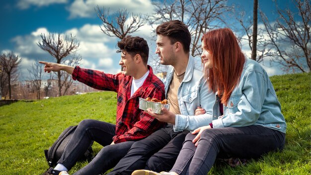 Three young friends sitting on the grass and holding food in a park