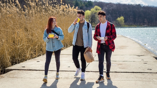 Three young friends walking along the lake, drinking and eating in a park