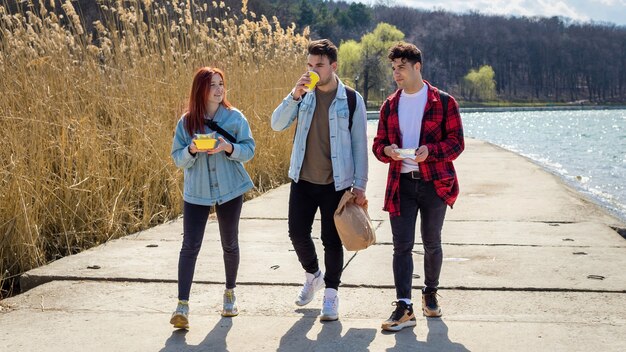 Three young friends walking along the lake, drinking and eating in a park