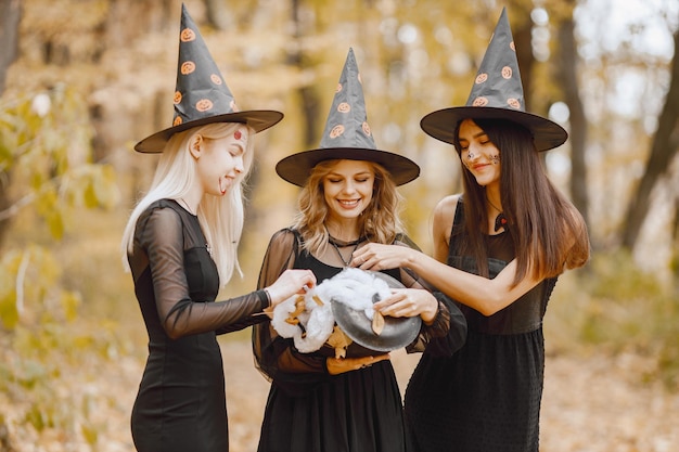 Foto gratuita tre giovani ragazze streghe nella foresta di halloween. ragazze che indossano abiti neri e cappello a cono. strega che tiene un calderone del mago