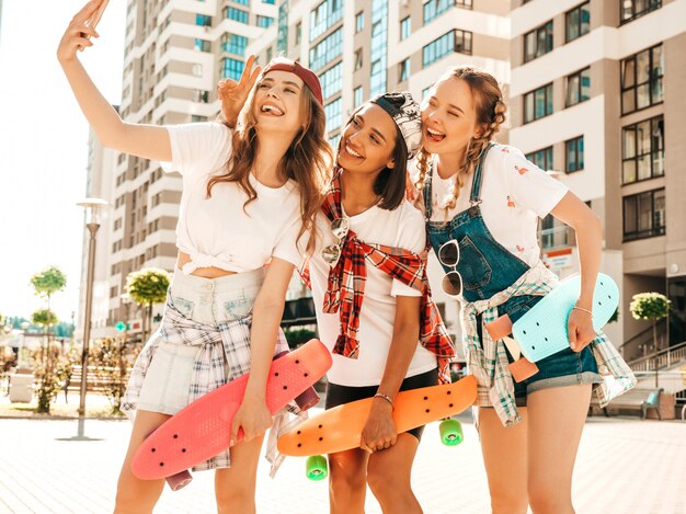 Три молодые улыбающиеся красивые девушки с красочными Пенни скейтборды. Женщины в летней одежде битник позирует на фоне улицы. Позитивные модели, принимая селфи автопортрет фотографии
