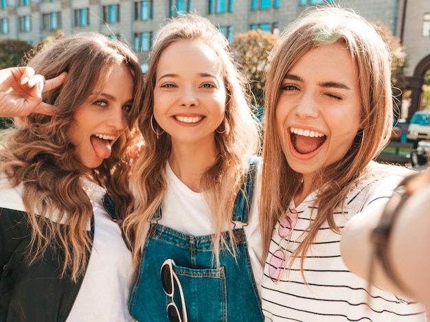 無料写真 夏服で3人の若い笑顔ヒップスター女性。スマートフォンでセルフポートレート写真を撮る女の子。通りでポーズをとるモデル。肯定的な顔の感情を示す女性