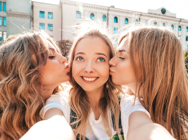 Foto gratuita tre giovani donne sorridenti dei pantaloni a vita bassa in vestiti di estate ragazze che prendono le foto dell'autoritratto del selfie sullo smartphone modelli che posano nella via femminile che bacia il loro amico in guancia