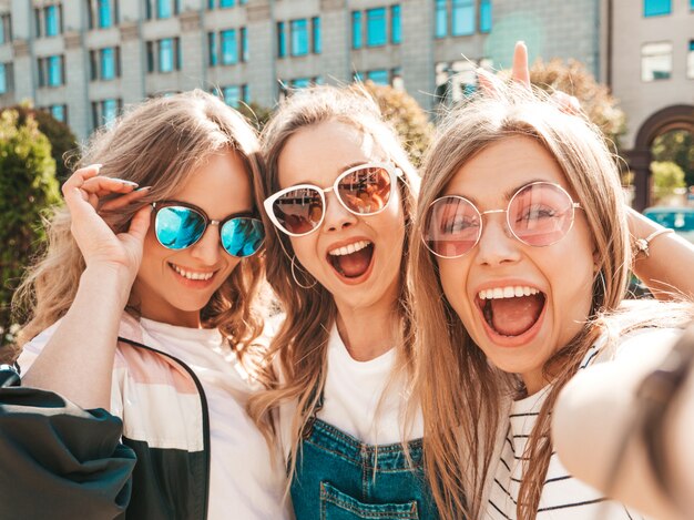 Три молодые улыбающиеся битник женщины в летней одежде. Девушки, делающие фотографии автопортрета селфи на смартфоне. Модели, позирующие на улице. Женщина, показывающая положительные эмоции лица в солнцезащитных очках