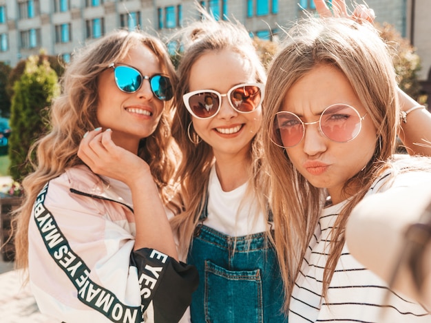 夏服で3人の若い笑顔の流行に敏感な女性。スマートフォンでセルフポートレート写真を撮る女の子。通りでポーズをとるモデル。サングラスで肯定的な顔の感情を示す女性