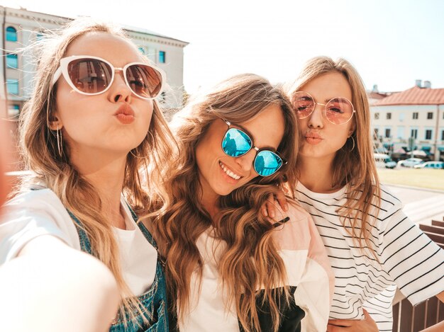 Три молодые улыбающиеся битник женщины в летней одежде. Девушки, делающие фотографии автопортрета селфи на смартфоне. Модели, позирующие на улице. Женщина, показывающая положительные эмоции лица в солнцезащитных очках