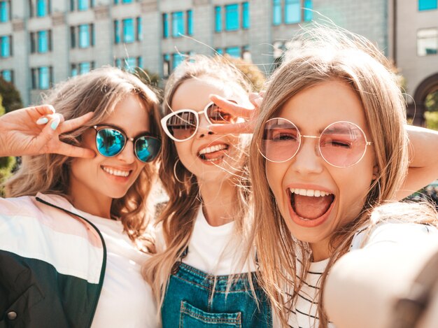 夏服で3人の若い笑顔ヒップスター女性。スマートフォンでセルフポートレート写真を撮る女の子。通りでポーズをとるモデル。肯定的な顔の感情を示す女性