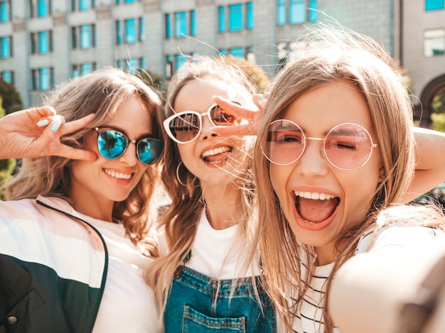 Three young smiling hipster women in summer clothes.Girls taking selfie self portrait photos on smartphone.Models posing in the street.Female showing positive face emotions