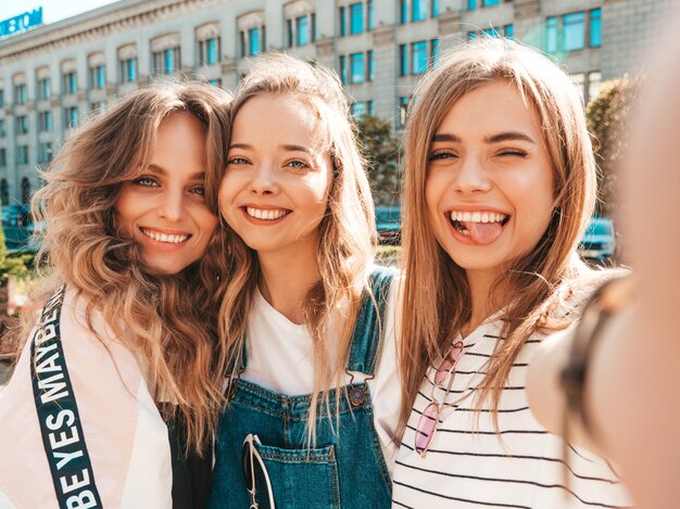 Три молодые улыбающиеся битник женщины в летней одежде. Девушки, делающие фотографии автопортрета селфи на смартфоне. Модели, позирующие на улице. Женщина, показывающая положительные эмоции лица