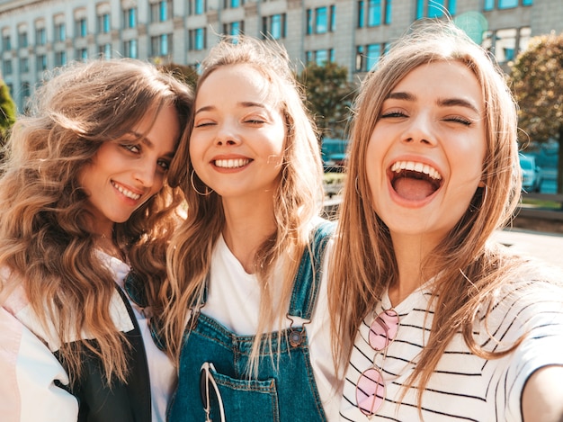 Three young smiling hipster women in summer clothes.Girls taking selfie self portrait photos on smartphone.Models posing in the street.Female showing positive face emotions