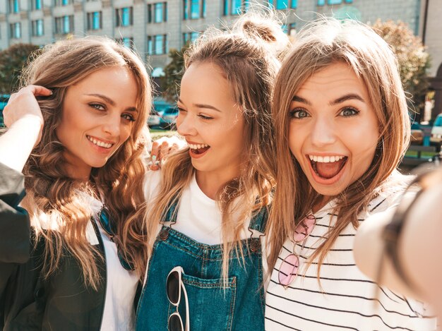 Three young smiling hipster women in summer clothes.Girls taking selfie self portrait photos on smartphone.Models posing in the street.Female showing positive face emotions