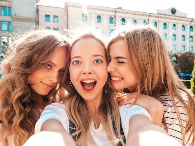 夏服で3人の若い笑顔ヒップスター女性。スマートフォンでセルフポートレート写真を撮る女の子。通りでポーズをとるモデル。肯定的な顔の感情を示す女性