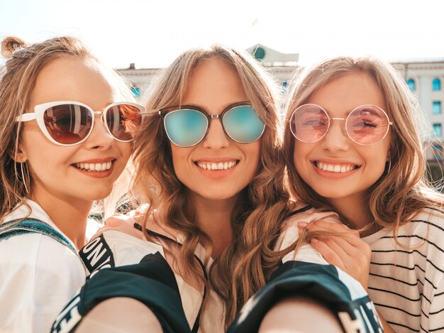 Three young smiling hipster women in summer clothes.Girls taking selfie self portrait photos on smartphone.Models posing in the street.Female showing positive face emotions