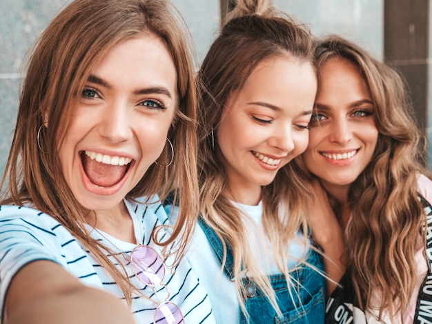 Три молодые улыбающиеся битник женщины в летней одежде. Девушки, делающие фотографии автопортрета селфи на смартфоне. Модели, позирующие на улице. Женщина, показывающая положительные эмоции лица