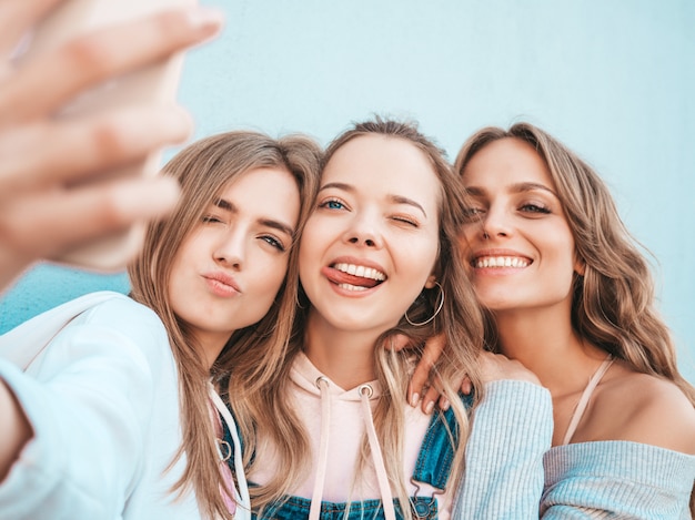 Три молодые улыбающиеся битник женщины в летней одежде. Девушки, делающие фотографии автопортрета селфи на смартфоне. Модели, позирующие на улице около стены. Женщина, показывающая положительные эмоции лица. Показывая язык