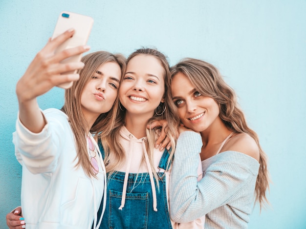 夏服の3人の若い笑顔の流行に敏感な女性。スマートフォンでselfieセルフポートレート写真を撮る女の子。壁の近くの通りでポーズをとるモデル。肯定的な顔の感情を示す女性