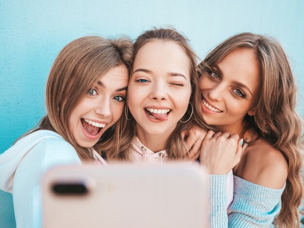 夏服の3人の若い笑顔の流行に敏感な女性。スマートフォンでselfieセルフポートレート写真を撮る女の子。壁の近くの通りでポーズをとるモデル。肯定的な顔の感情を示す女性