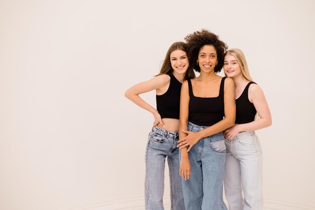 three young smiling intercultural women wearing top and jeans looking at camera on white background with space for text