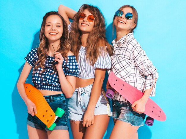 Three young stylish smiling beautiful girls with colorful penny skateboards. Women in summer clothes posing in sunglasses. Positive models having fun