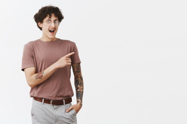 Thrilled and amazed happy hipset guy with curly hairstyle stylish moustache and tattoos gasping smiling joyfully and staring astonished while pointing at upper right corner holding hand in pocket