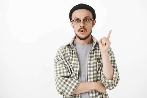 Thrilled and amused stylish male in black beanie and glasses raising index finger in eureka gesture
