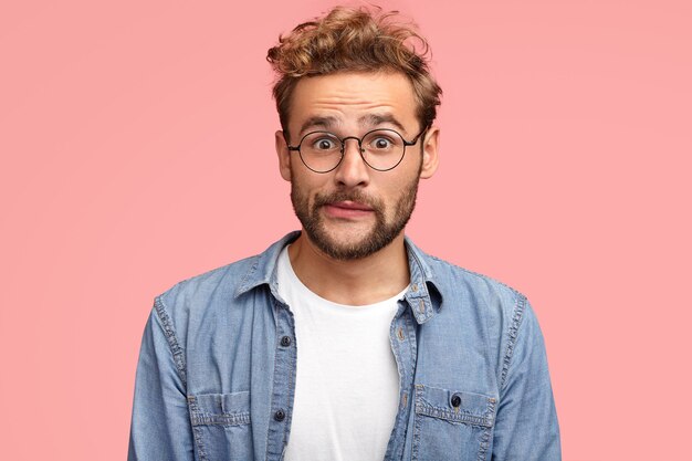 Thrilled curly hipster with puzzled expression purses lips and looks with stunned expression, wears denim shirt, has stubble, isolated over pink wall. People, reaction and lifestyle concept