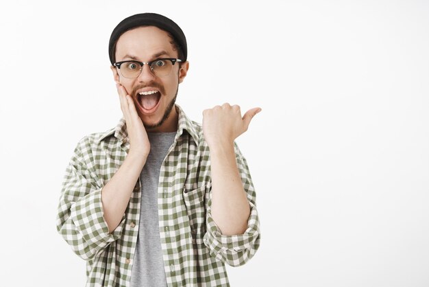 Thrilled excited and amazed good-looking young male device user in black beanie green shirt pointing right with astonished and impressed expression holding hand on face and dropping jaw