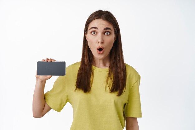 Thrilled girl shows horizontal smartphone screen, gasp and say wow, showing application on mobile phone, interface of app on cellphone, standing on white.