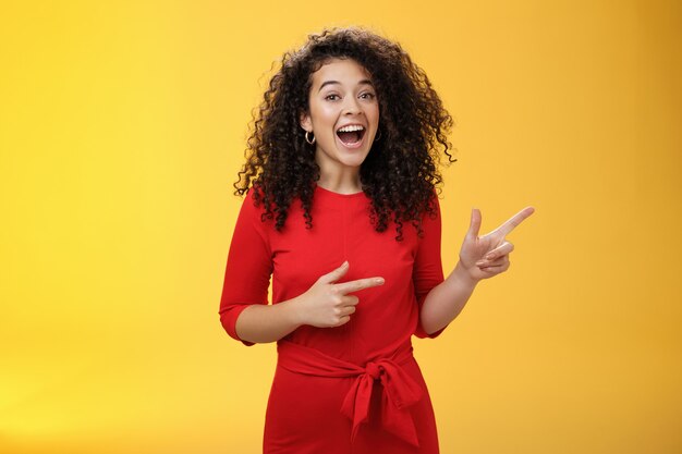 Thrilled and happy joyful young charismatic woman with curly hair in red dress laughing out loud and smiling amazed, delighted pointing at upper right corner impressed over yellow background.