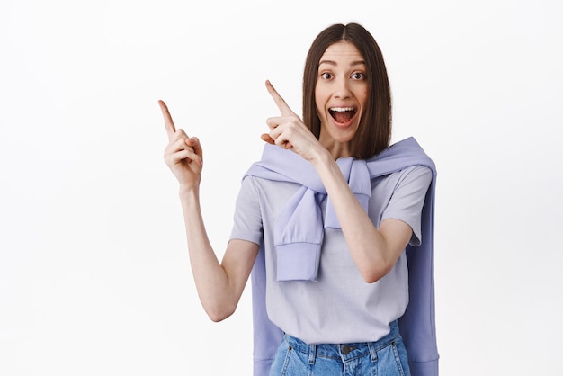 Thrilled happy woman pointing at upper left corner looking amazed while showing product on discount indicating at banner standing over white background