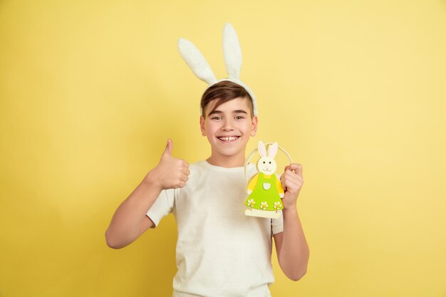 Thumb up. Decorating. Caucasian boy as an Easter bunny on yellow studio background. Happy easter greetings. Beautiful male model. Concept of human emotions, facial expression, holidays. Copyspace.