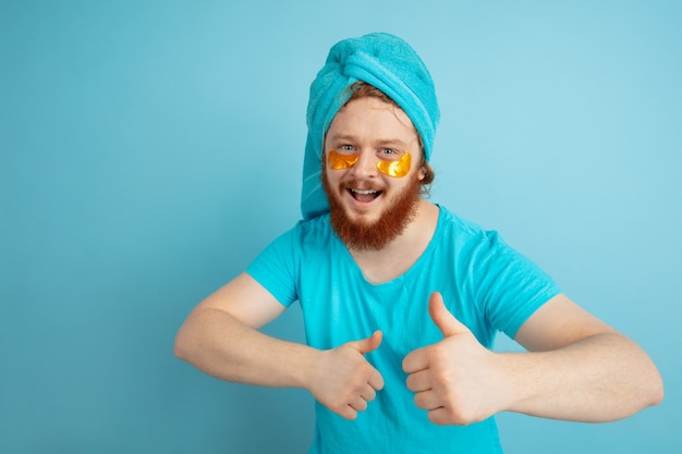 Thumb up. Portrait of young caucasian man in his beauty day and skin care routine. Male model with natural red hair applying under-eye golden patches. Body and face care, natural, male beauty concept.