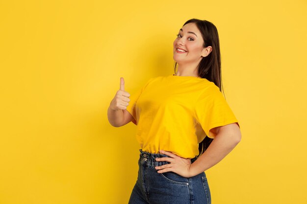 Thumb up, smiling. Caucasian woman's portrait isolated on yellow wall.