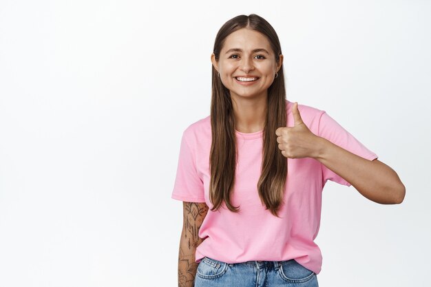 Thumbs up. Smiling woman in pink t-shirt shows her support, recommending product, like and agree, nod in approval, standing on white.