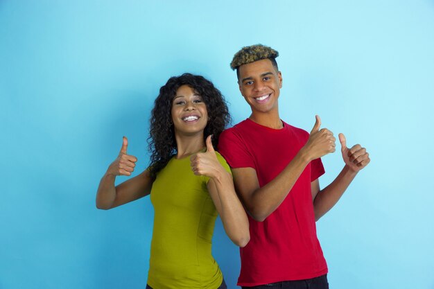 親指を立てて、笑顔。青の背景にカラフルな服を着た若い感情的なアフリカ系アメリカ人の男性と女性。美しいカップル。人間の感情、顔の表現、関係、広告、友情の概念。