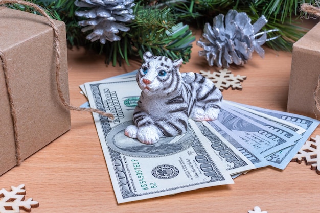 The tiger is the symbol of the chinese new year 2022. toy tiger holding cash dollars on a wooden background with gift boxes, snowflakes and fir branches with cones. success and wealth concept in 2022