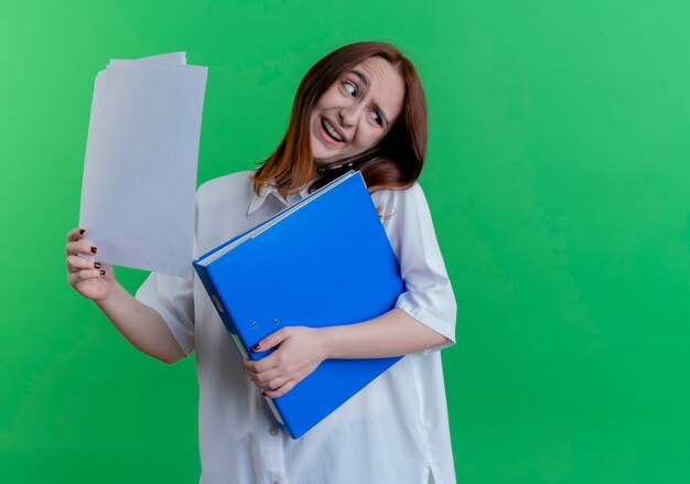 Tilting head smiling young redhead girl holding paper with folder and speaks on phone isolated on green