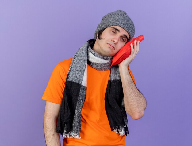 tilting head young ill man wearing winter hat with scarf putting hot water bag on cheek isolated on purple