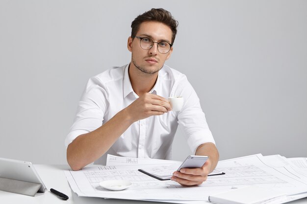 Time for break. Job, relaxation and modern electronic devices. Successful professional chief engineer browsing internet on mobile phone and enjoying espresso, having rest during working day at office