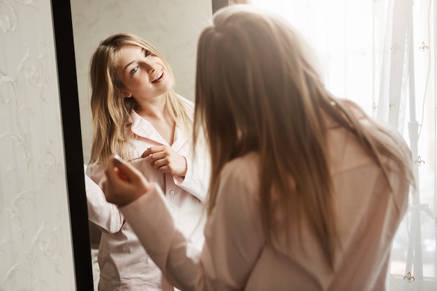 Time to dress up and went to meet adventures. Home shot of beautiful blond caucasian girl looking in mirror, wearing nightwear and touching hair strand, thinking about new hairstyle