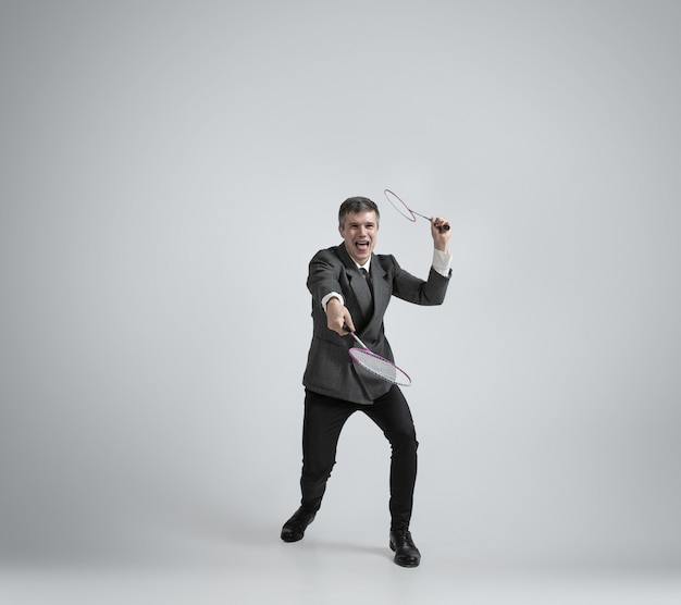 Time for emotions. Man in office clothes plays badminton with two racket on grey background