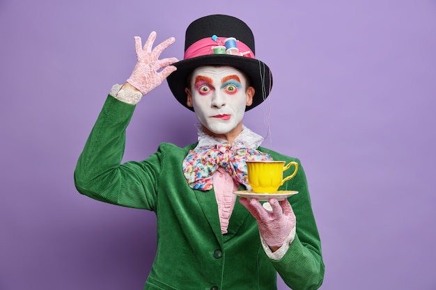 Time for having tea. Aristocratic gentleman with bright makeup has image of fictional character holds cup of drink wears big hat has wondered expression poses over purple wall