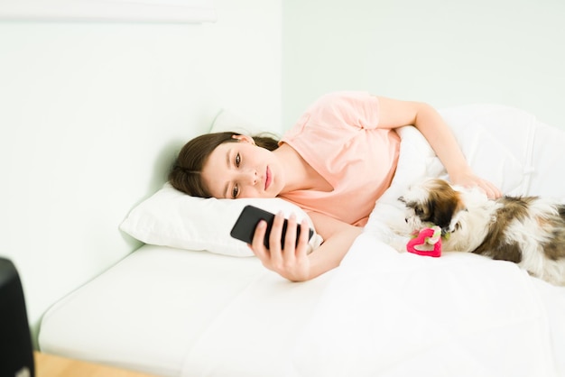 Time to sleep. Tired young woman texting and looking at social media on her smartphone while trying to fall asleep with her cute shih tzu puppy