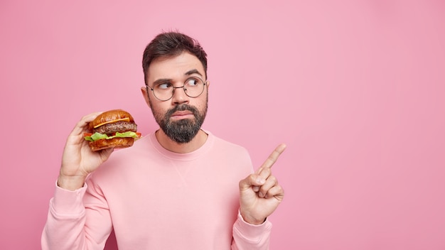 Time for snack. Serious bearded adult man holds delicious burger eats cheat meal points on blank space wears round spectacles casual clothes