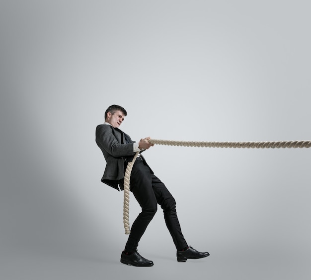 Free photo time for strenght. man in office clothes training with ropes on grey wall. get target, overcoming problems, deadlines. businessman in motion, action. sport, healthy lifestyle, working.