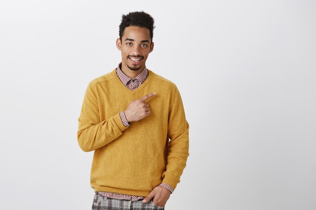 Time to visit new places. Portrait of charming young dark-skinned guy with afro haircut smiling politely and pointing at upper right corner with index finger, smiling while promoting copy space