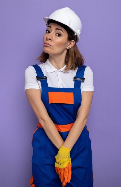 Foto gratuita timido giovane donna costruttore in uniforme da costruzione e casco di sicurezza in guanti di gomma con le mani insieme in piedi sul muro viola