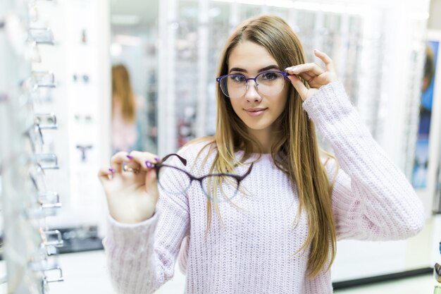 白いセーターを着た小さな女性が腕に医療用メガネを持ち、特別な店でそれらを見る
