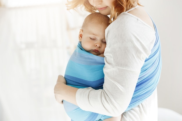 Tiny newborn child close eyes and having good sleep in baby sling feeling protection from his beautiful young mother. Family, lifestyle concept.