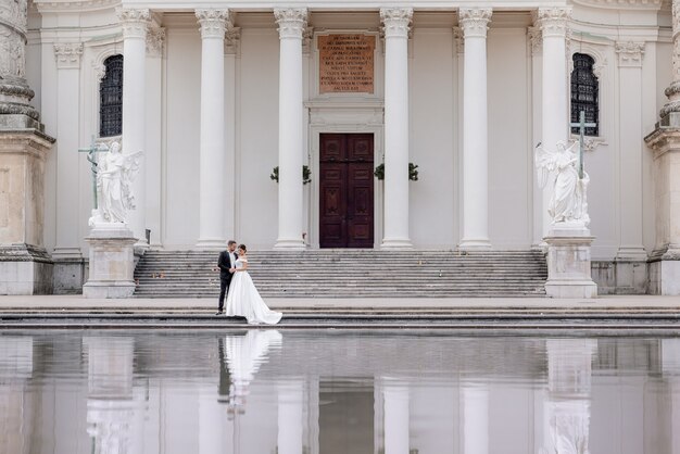 작은 웨딩 커플은 흰색 열과 물에 반사와 거대한 성당 근처에 걷고있다